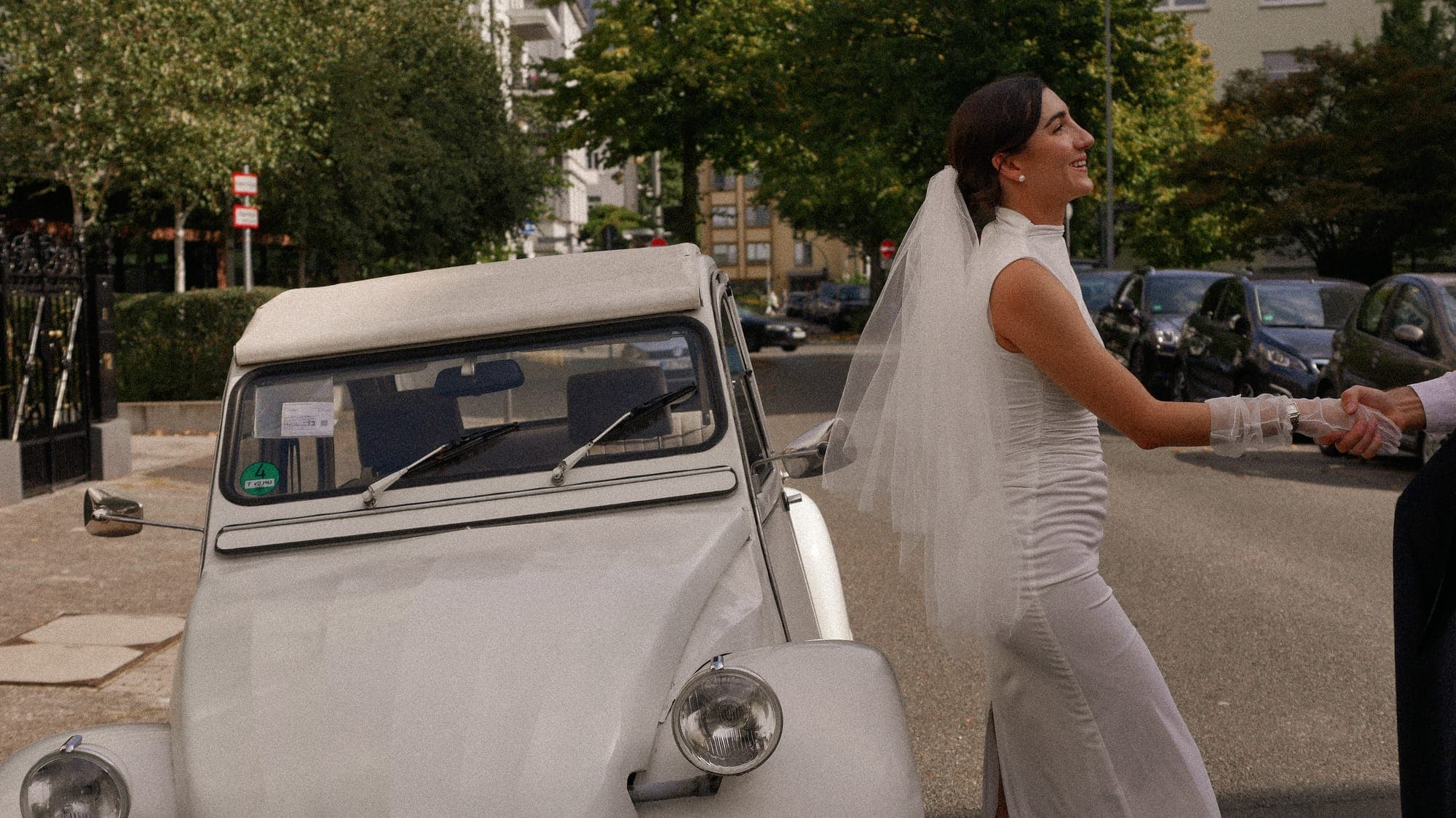 Braut winkt lachend neben einem alten Citroën 2CV
